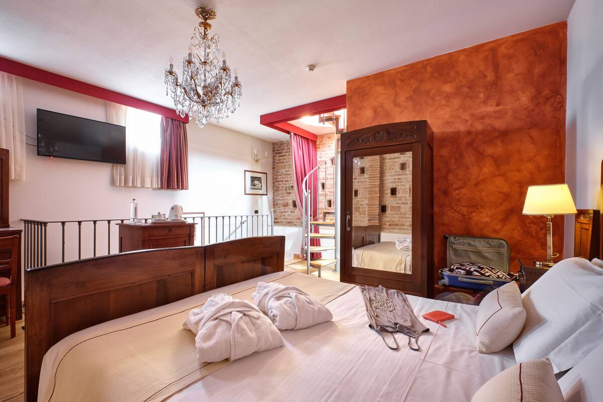 Une chambre d’hôtel dotée d’un grand lit, d’une tête de lit en bois, d’un lustre, d’une armoire, d’une télévision et d’un escalier menant à un espace extérieur.
