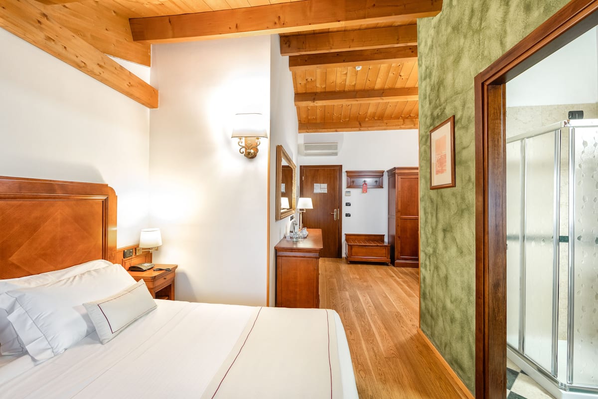 Une chambre d’hôtel avec un plafond en bois, un lit avec du linge de maison blanc, une table de chevet en bois, un bureau avec une lampe, un miroir et une salle de bain avec une douche.