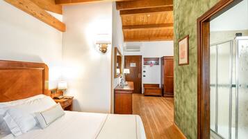 Ein Hotelzimmer mit Holzboden, einem Bett mit weißen Leinentüchern, einem Nachttisch aus Holz, einem Schreibtisch mit Lampe, einem Spiegel und einem Badezimmer mit Dusche.