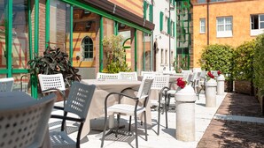 An outdoor cafe with tables and chairs, a brick building with green shutters, and a cobblestone path. - Hotel Villa Malaspina (Castel d'Azzano)