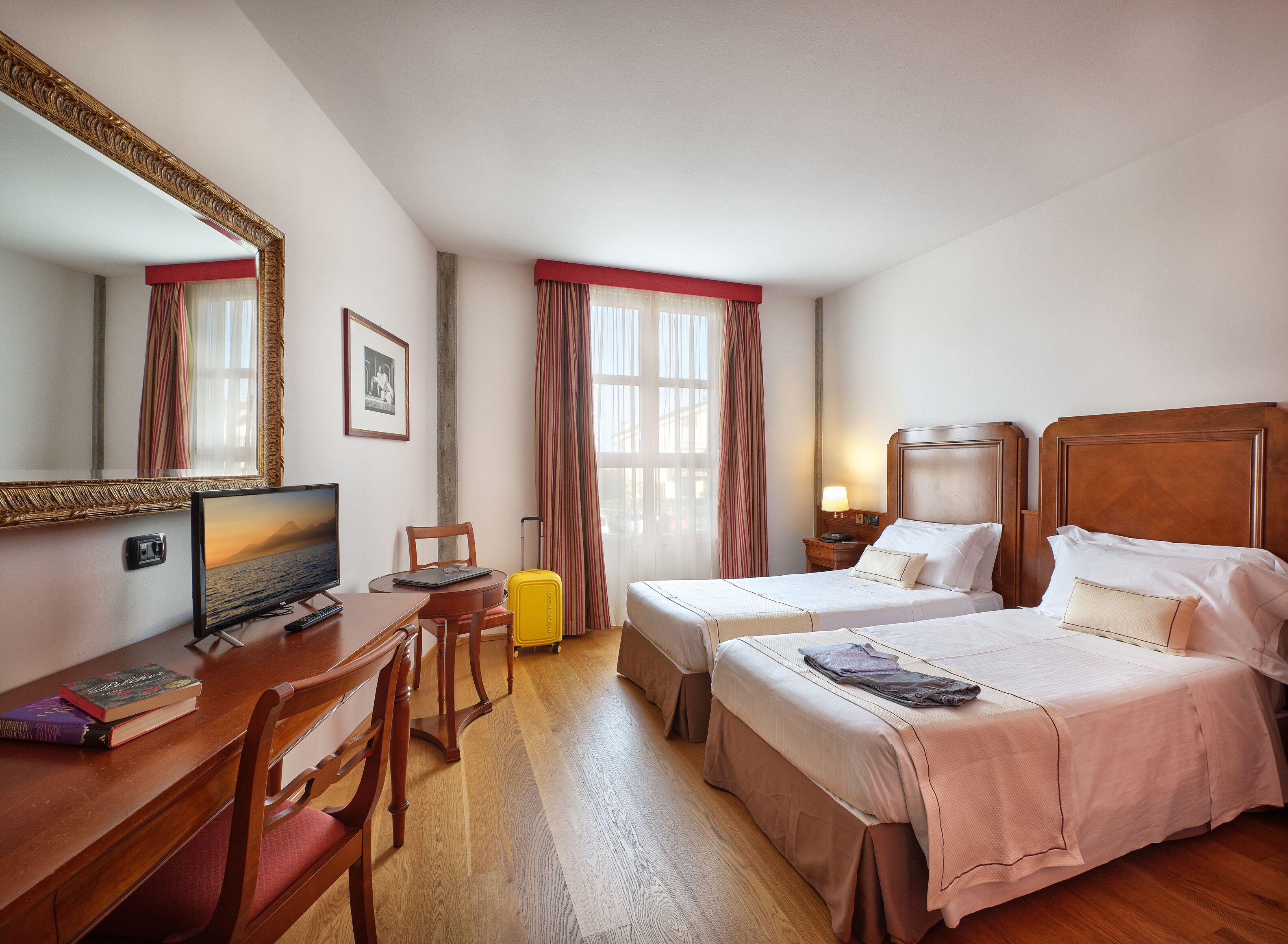 Une chambre d’hôtel équipée d’un bureau, de deux lits, d’une chaise, d’une valise et d’un grand miroir.
