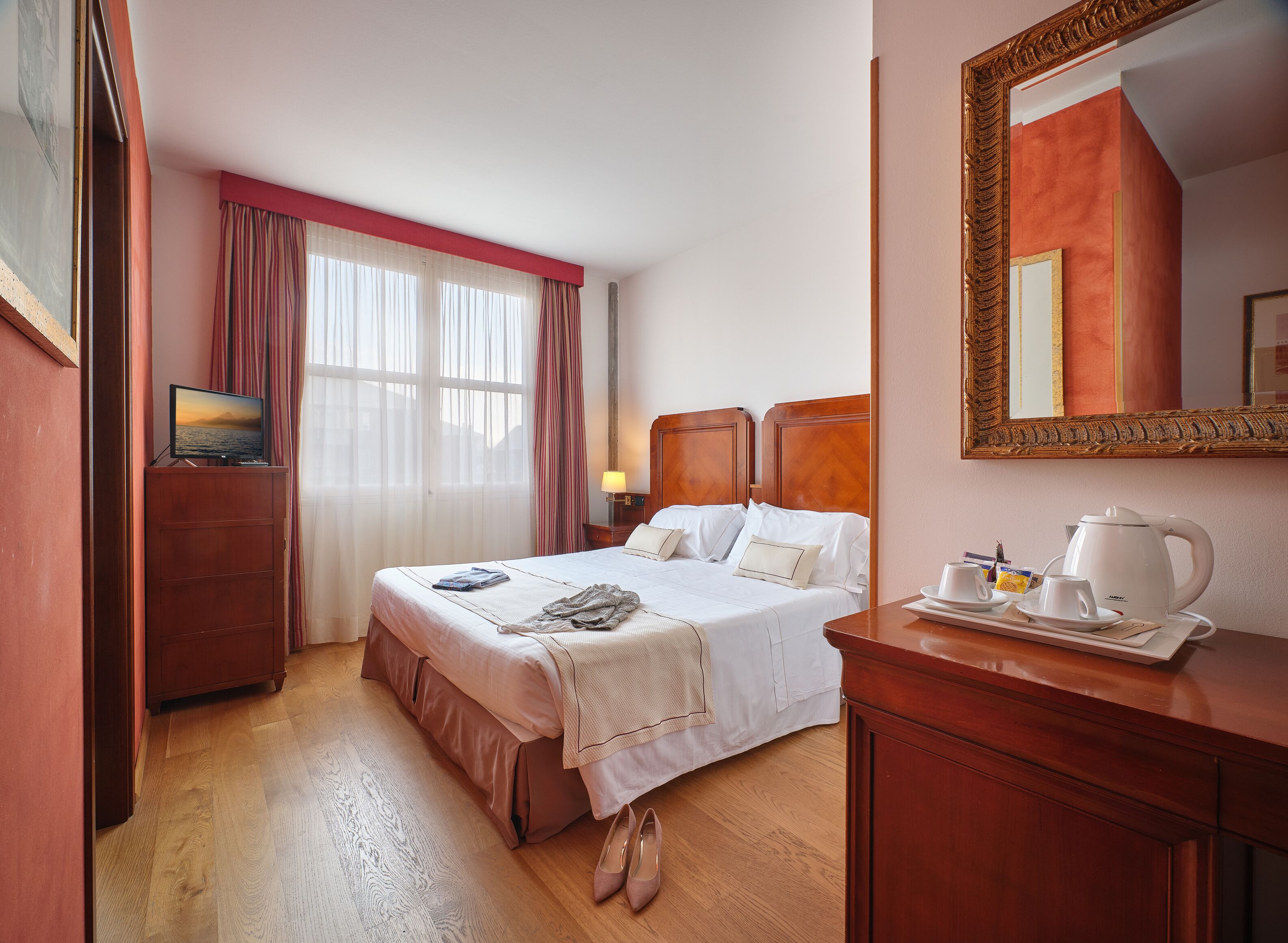 Habitación de hotel con cama, cómoda de madera, espejo y ventana con cortinas.