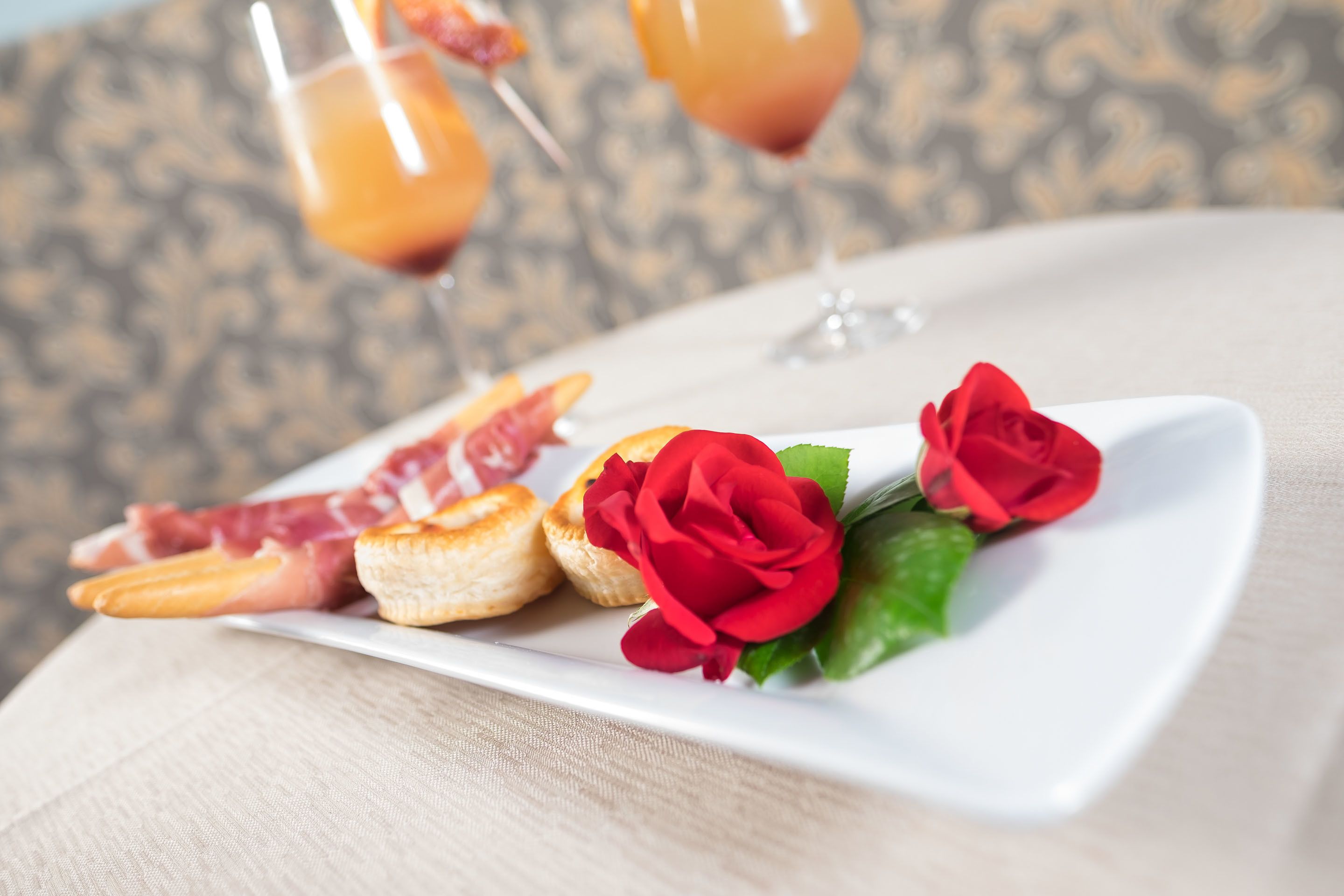 A white rectangular plate with pastries, a red rose, and two glasses of orange beverage on a table.