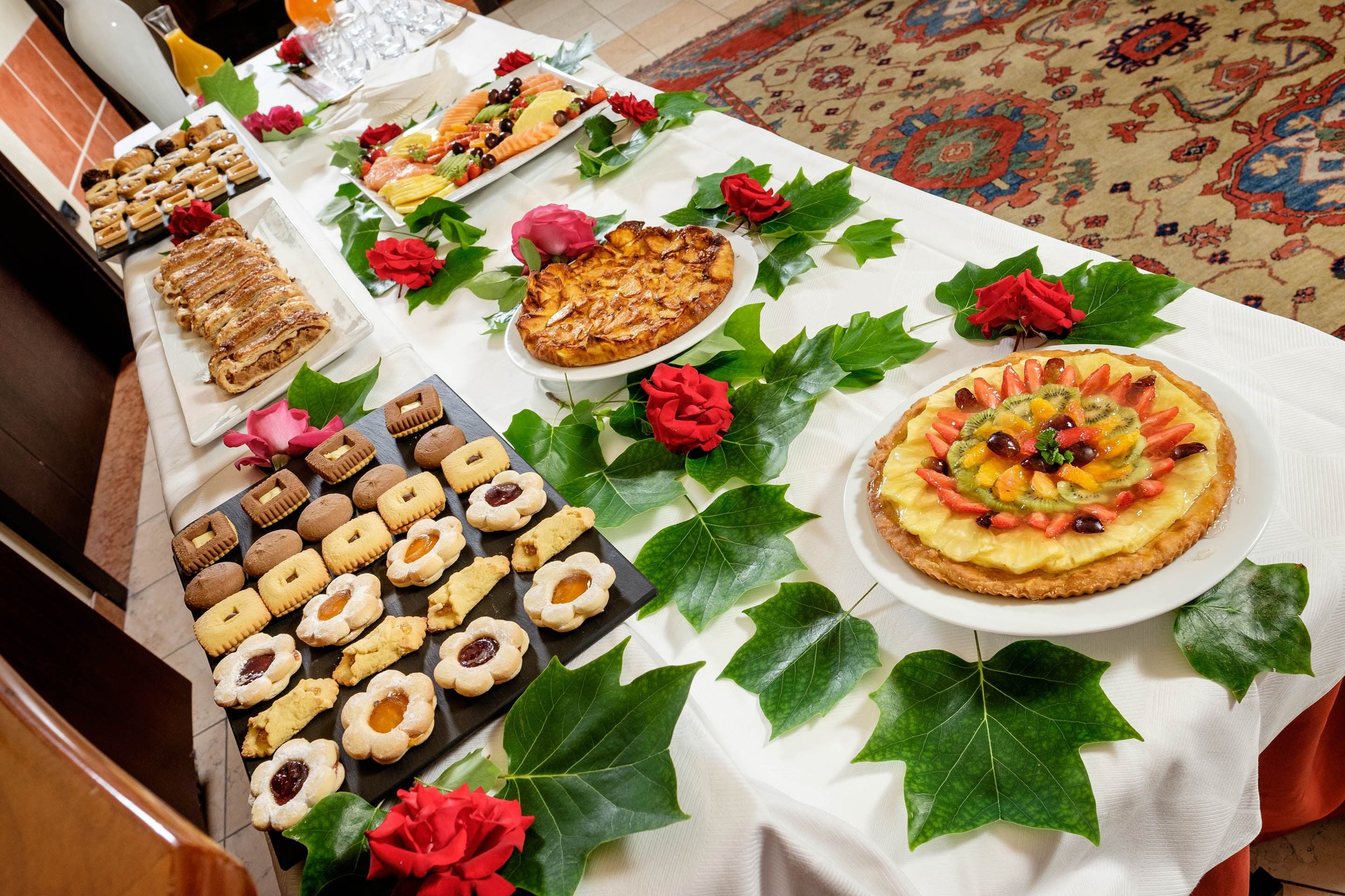 A table with various pastries and desserts, including tarts and cookies, arranged on a white tablecloth with green leaves and red flowers.