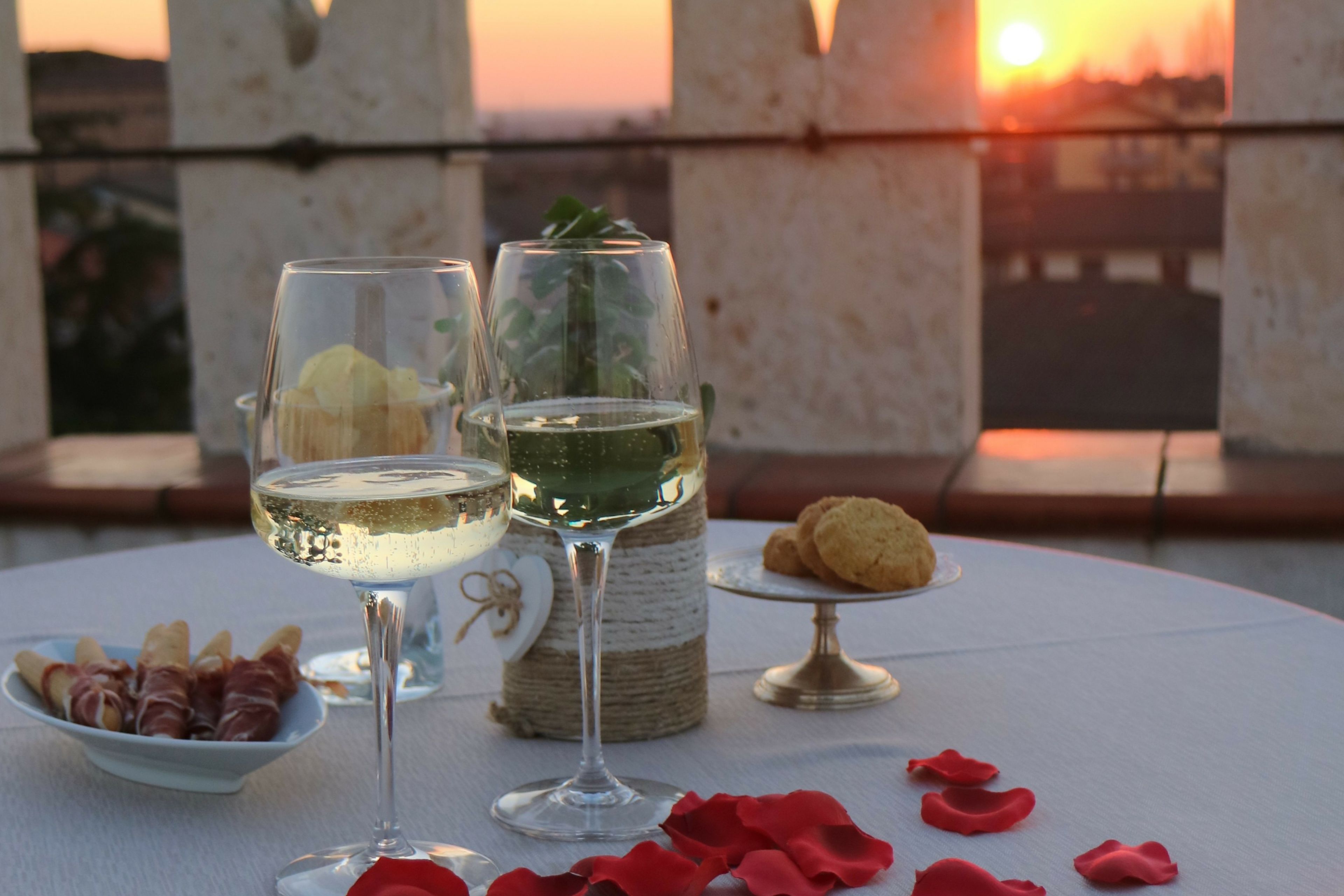 A table with two glasses of white wine, a small bowl of prosciutto, a plate of cookies, and red rose petals.