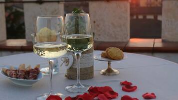 A table with two glasses of white wine, a small bowl of prosciutto, a plate of cookies, and red rose petals.