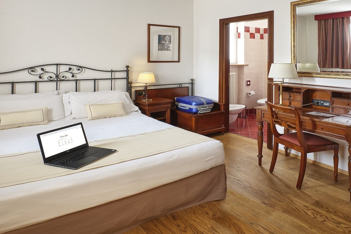 Une chambre d’hôtel avec un lit, un bureau et une chaise, un ordinateur portable, une valise et un tableau encadré au mur.