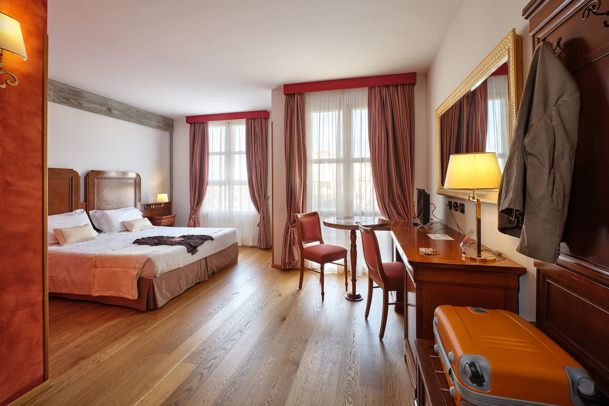 Une chambre d’hôtel avec un lit, un bureau, deux chaises, une table, un miroir et une valise.