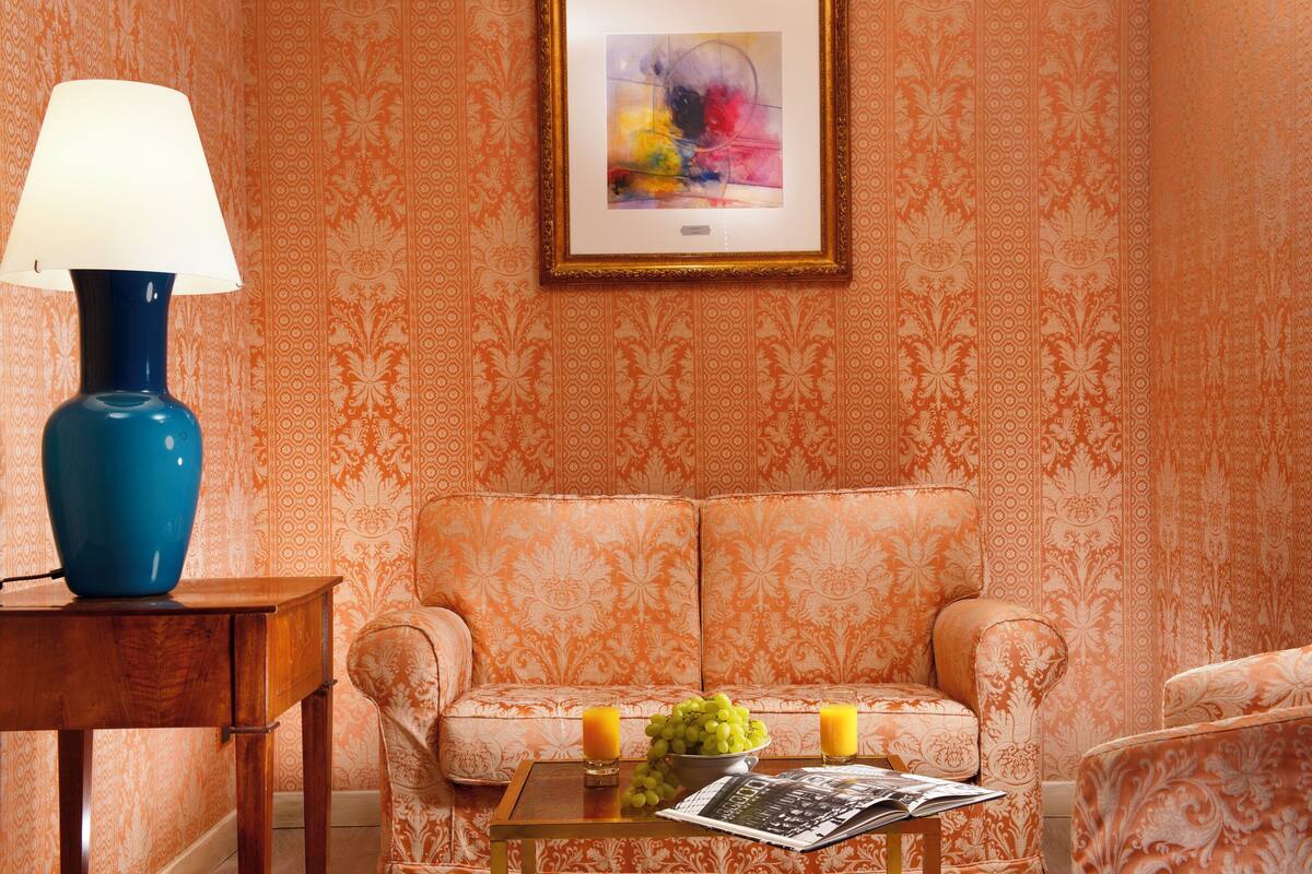 Ein Zimmer mit gemustertem Wandpapier, einer blauen Lampe auf einem Holztisch, einer Couch mit einer Zeitschrift und Obst, sowie einem gerahmten abstrakten Gemälde.