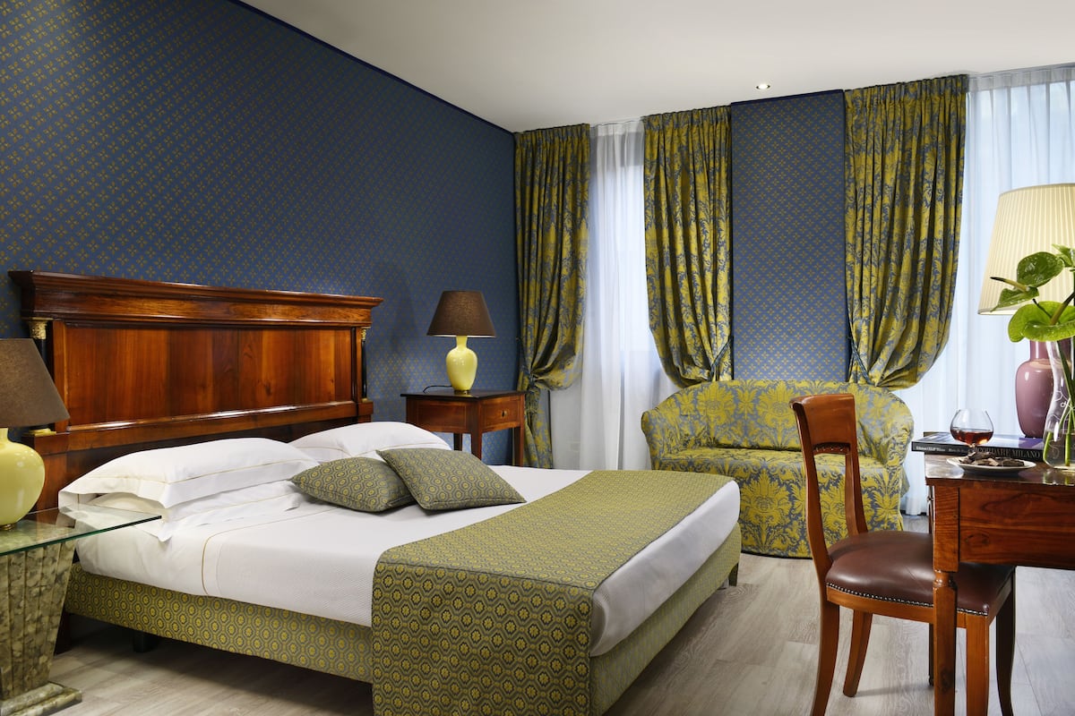 Ein Hotelzimmer mit einem hölzernen Bett, einer grün gemusterten Tagesdecke, einem Nachttisch aus Holz, einer Lampe, einem Sessel, einem Tisch mit einer Vase und Vorhängen.