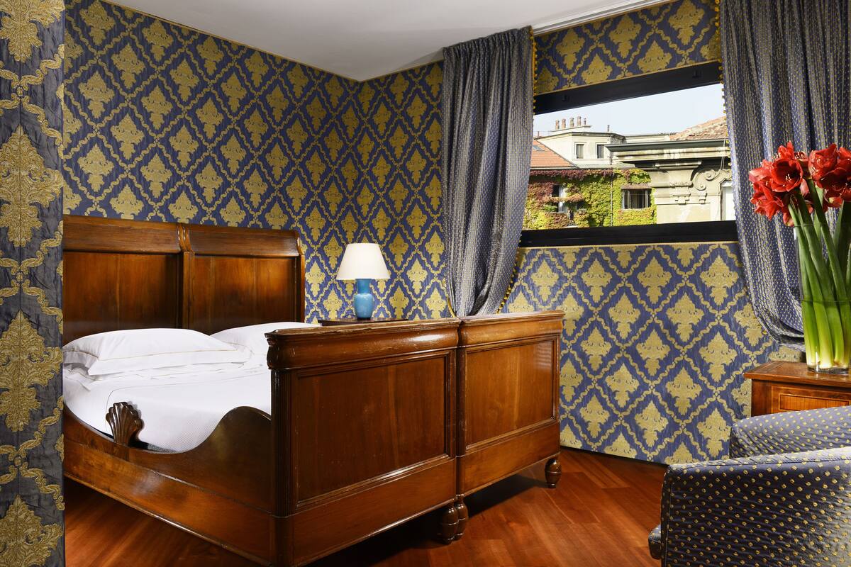 Ein Schlafzimmer mit einem hölzernen Bett, einem Nachttisch mit Lampe, einem Fenster mit Blick auf Gebäude und Wandpapier mit Muster.
