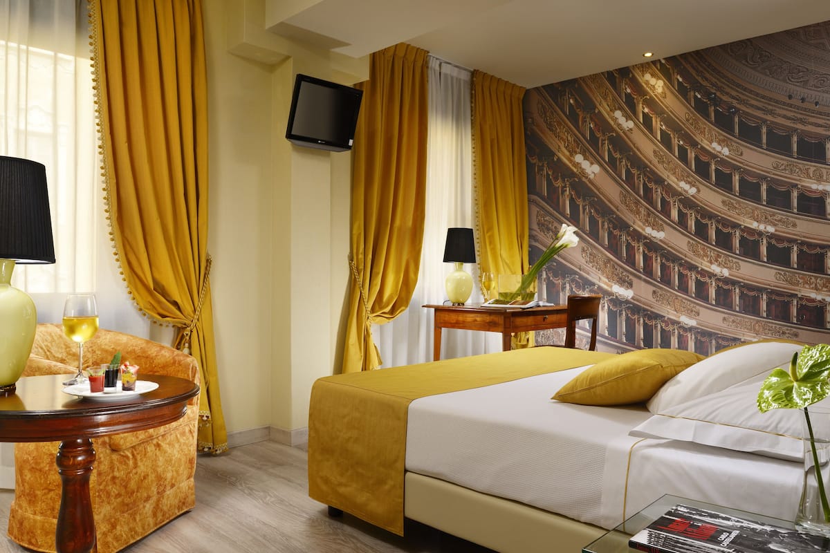 Ein Hotelzimmer mit Bett, einer Couch, einem Tisch mit Lampe, einem Fernseher und einem Wandgemälde eines Opernhauses.