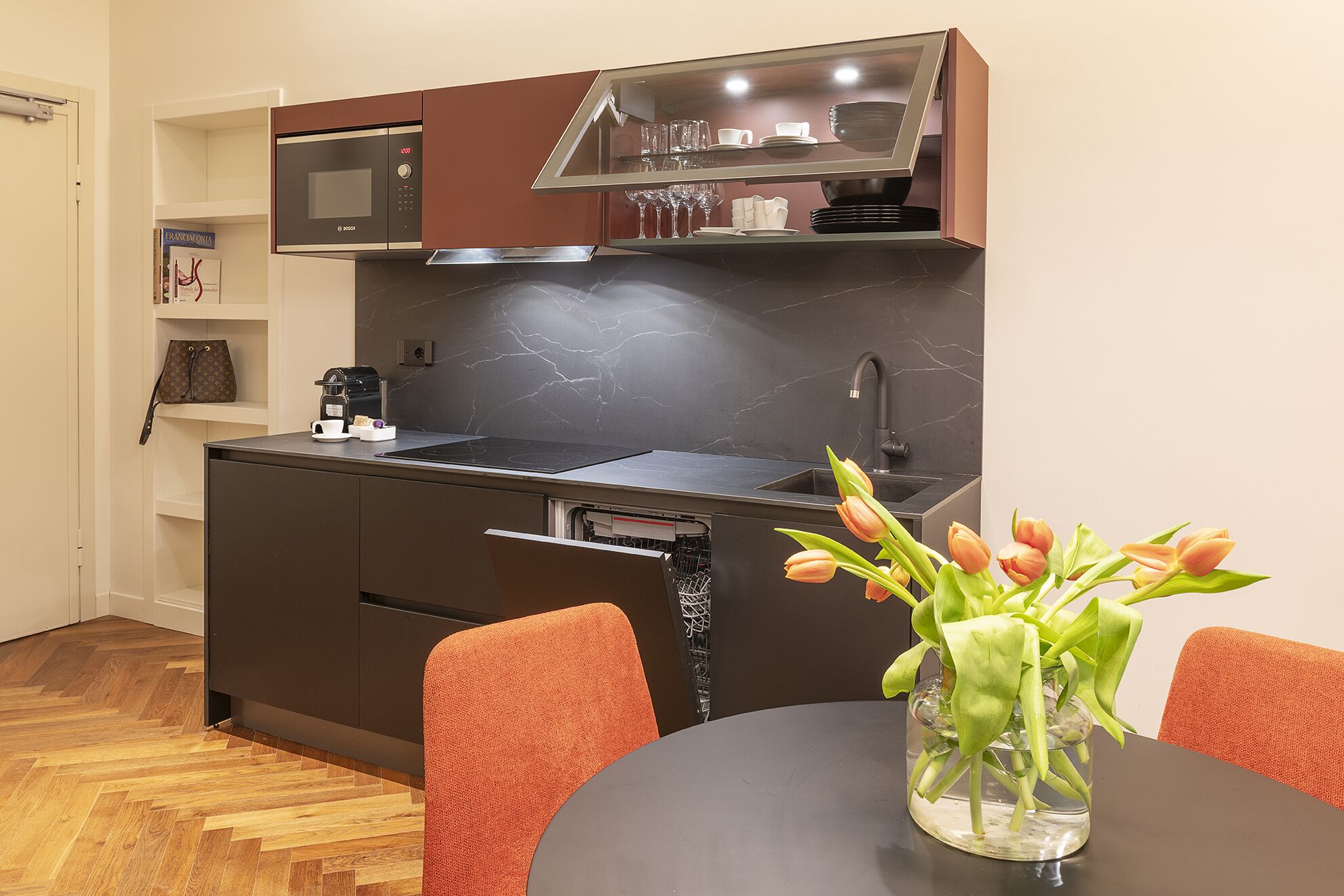 Cocina pequeña con microondas, pileta y una pequeña zona de encimera. Hay una mesa de comedor con sillas y un florero con flores.