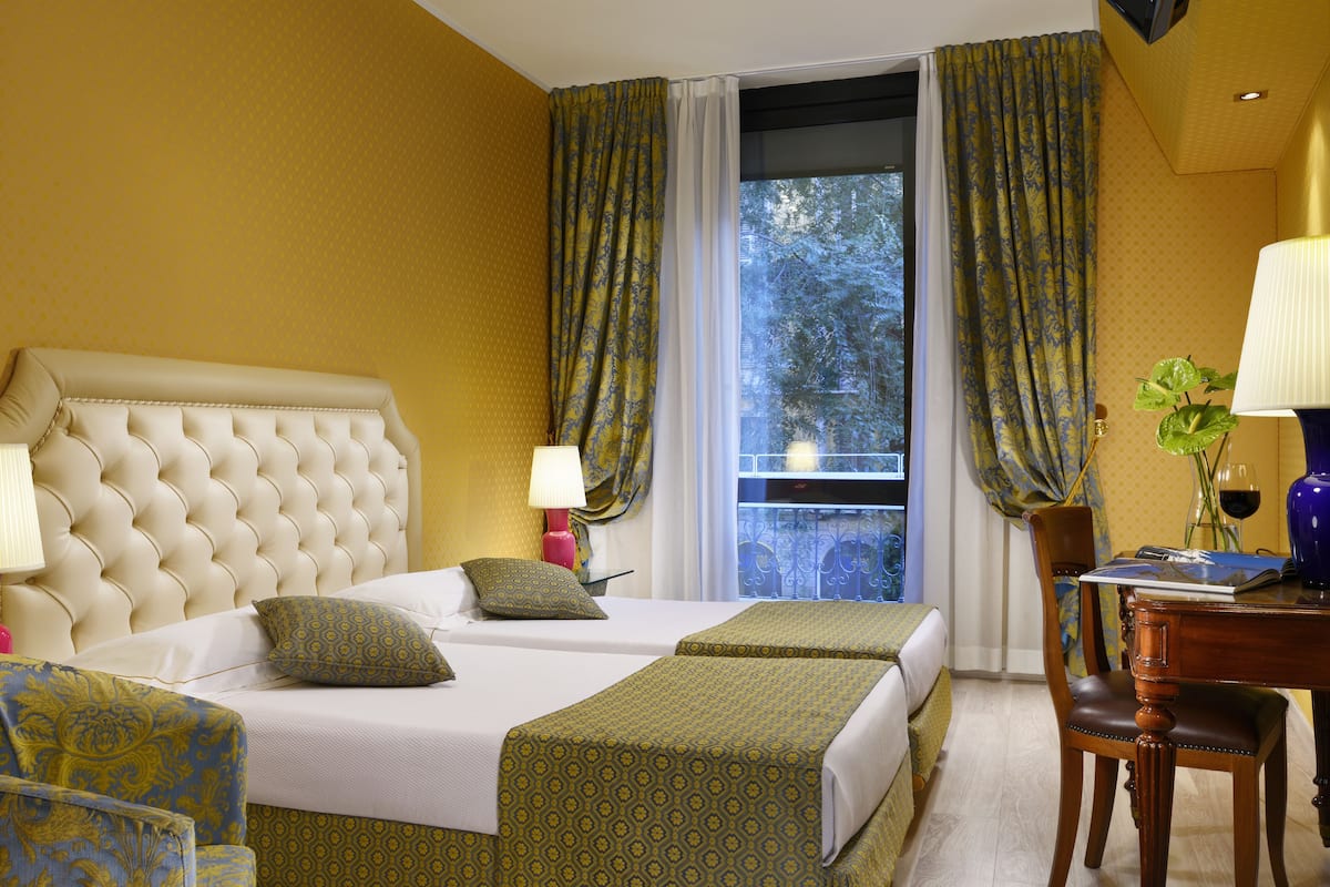 Ein Hotelzimmer mit einem großen Bett, zwei Nachttischen, einem Schreibtisch mit Stuhl und einem fensterbepflanzten Fenster.