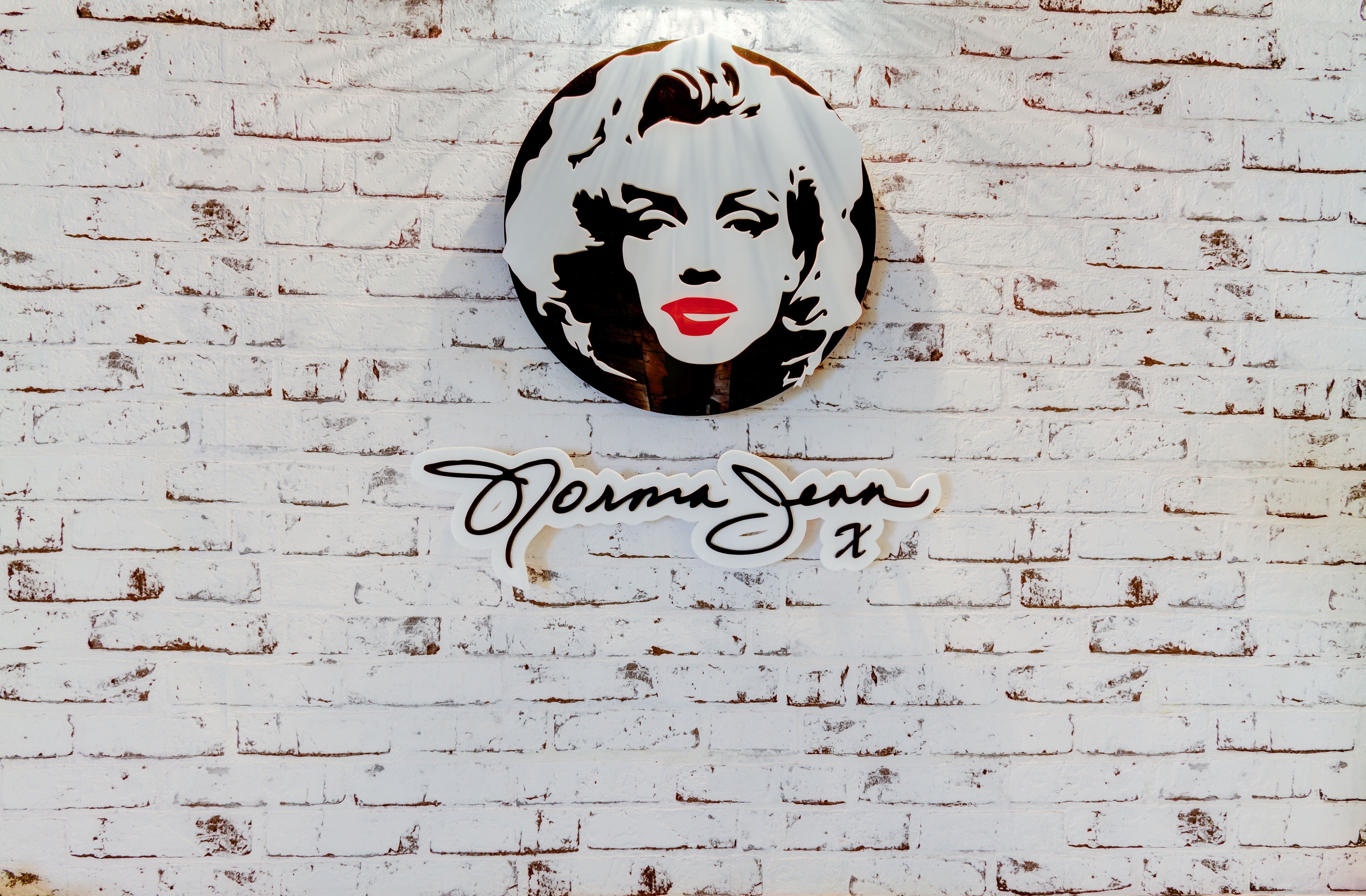 Un mur en briques blanches sur lequel est peinte une œuvre d’art circulaire représentant un portrait stylisé, avec le nom « Norma Jean » inscrit en dessous.