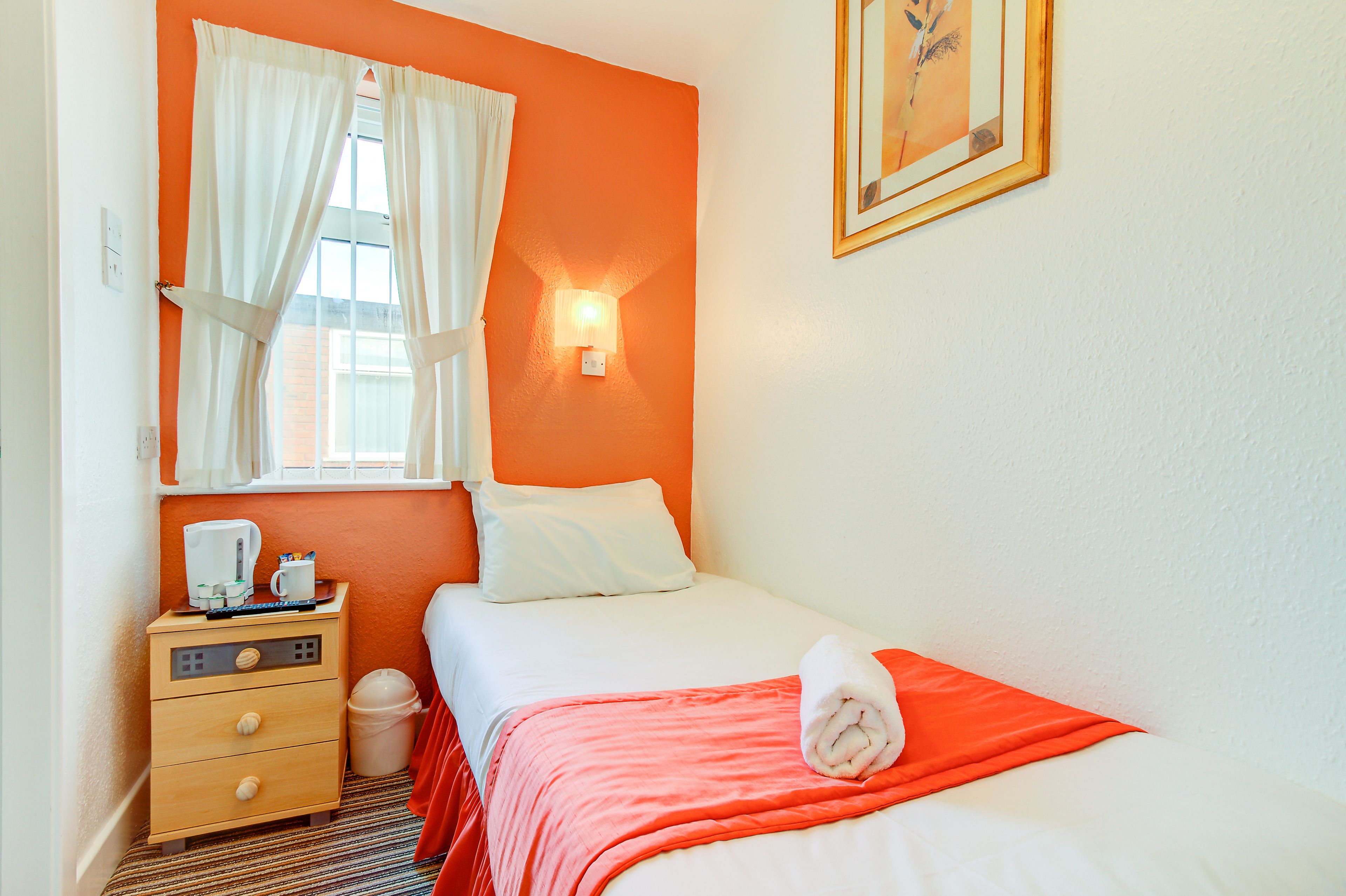 A hotel room with a bed, a nightstand, a window with curtains, a wall-mounted light, and a framed picture on the wall.