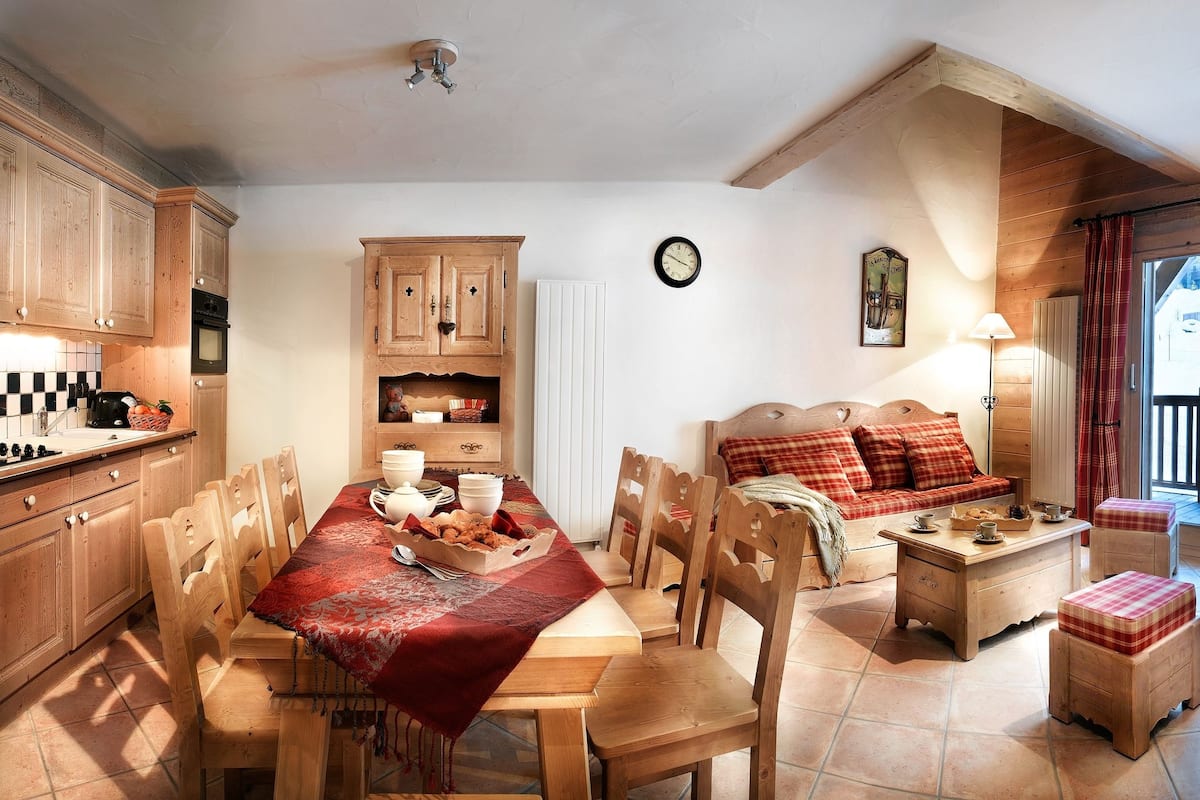 A cozy kitchen with wooden cabinets, a dining table with chairs, and a red tablecloth. There is a clock on the wall and a television in the corner.
