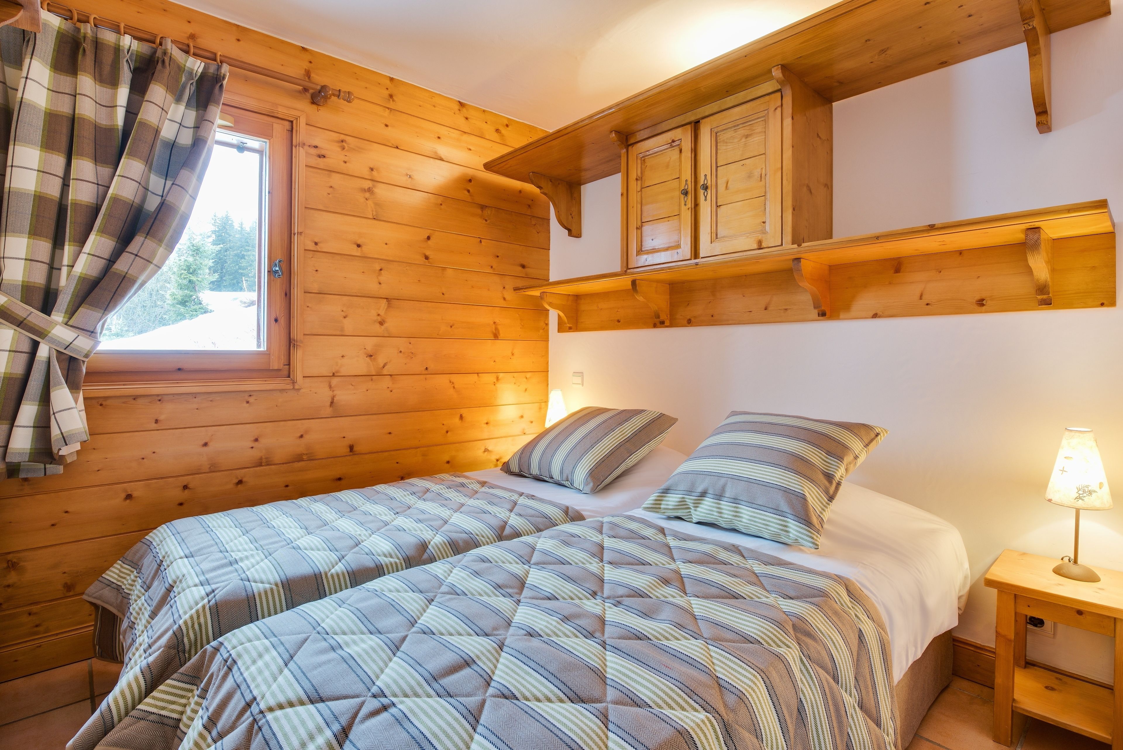 A wooden cabin room with two beds, a window with curtains, a wooden cabinet, and a bedside lamp.
