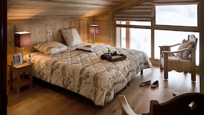 A cozy wooden cabin bedroom with a large bed, bedside lamps, a chair, and a view through the window. - CGH Résidences & Spas Les Cimes Blanches (Montvalezan)