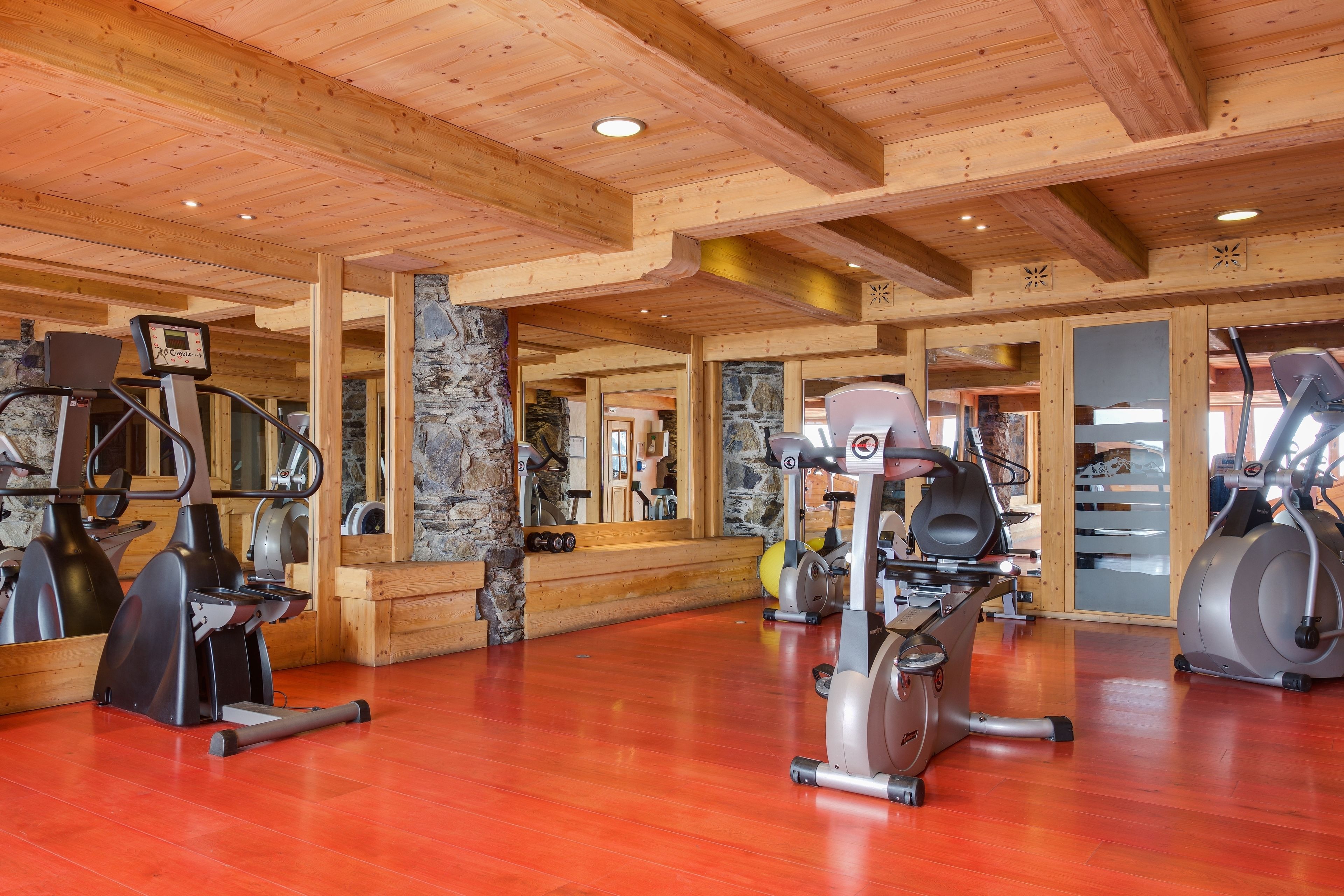 A gym with various exercise equipment, wooden beams on the ceiling, and stone walls.