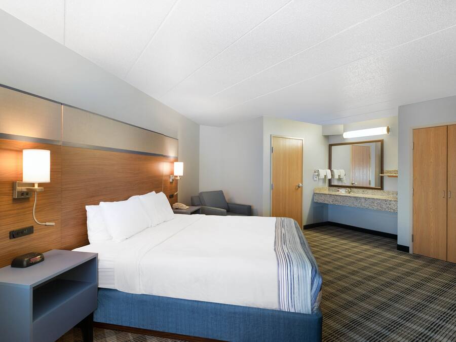 A hotel room with a bed, bedside table, lamp, chair, and a bathroom with a mirror and sink.