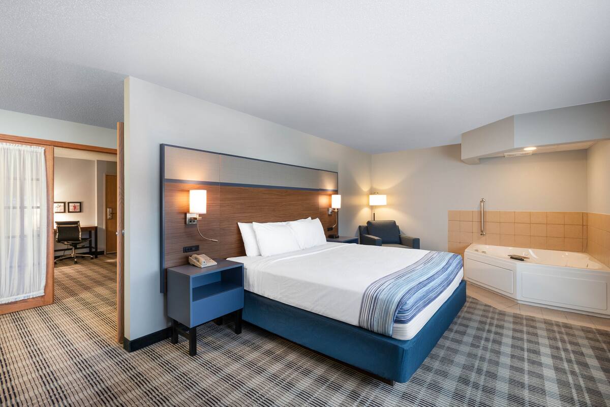 Habitación de hotel con una cama grande, un escritorio con silla, una lámpara de escritorio, una mesita de noche, un teléfono, una luz en la pared, un baño con bañera y ducha.