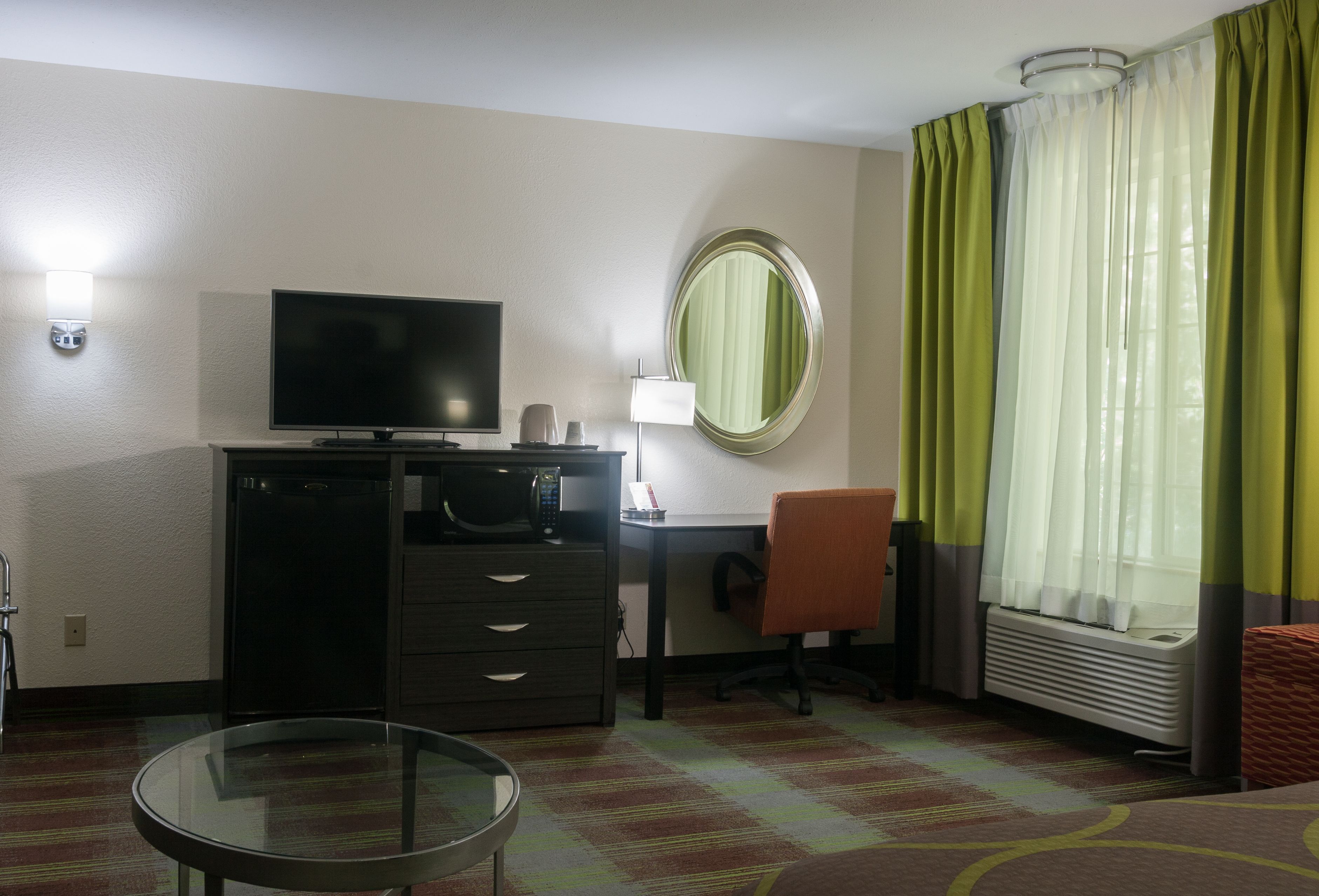 Une chambre d’hôtel équipée d’un téléviseur à écran plat, d’un bureau, d’une chaise, d’une petite table, d’un miroir et d’une fenêtre avec des rideaux.