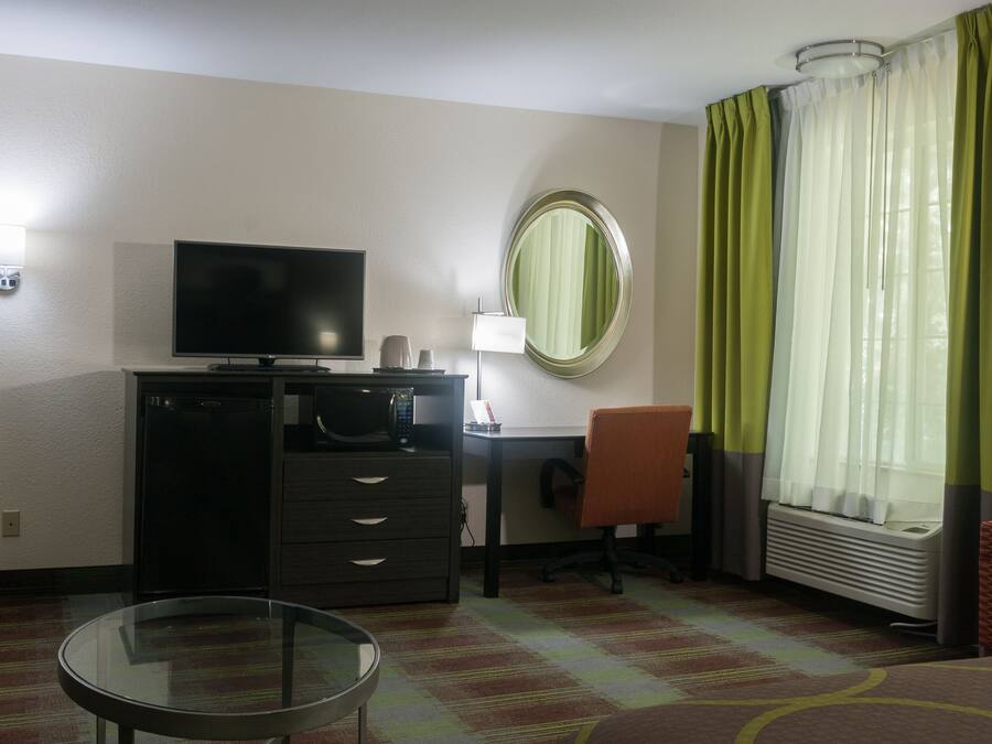 Une chambre d’hôtel équipée d’un téléviseur à écran plat, d’un bureau, d’une chaise, d’une petite table, d’un miroir et d’une fenêtre avec des rideaux.