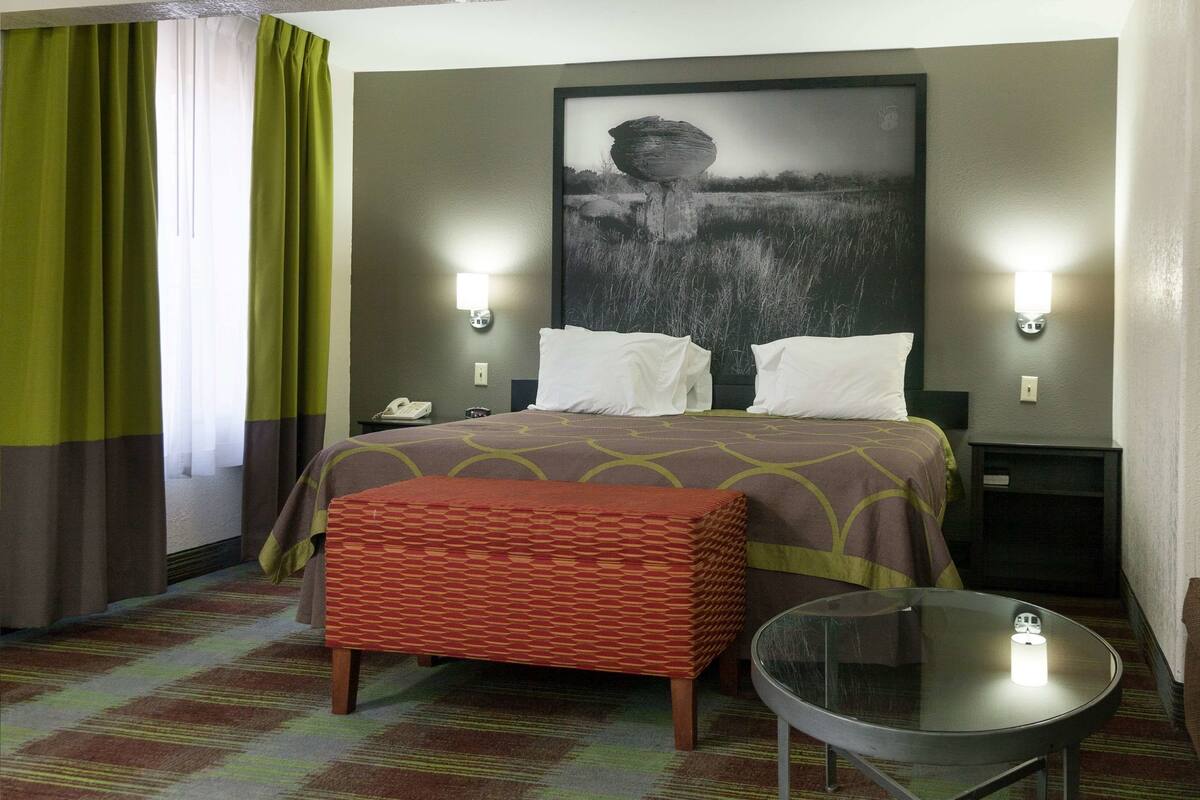 A hotel room with a bed, two bedside tables, a glass table, a patterned ottoman, and a large framed picture on the wall.