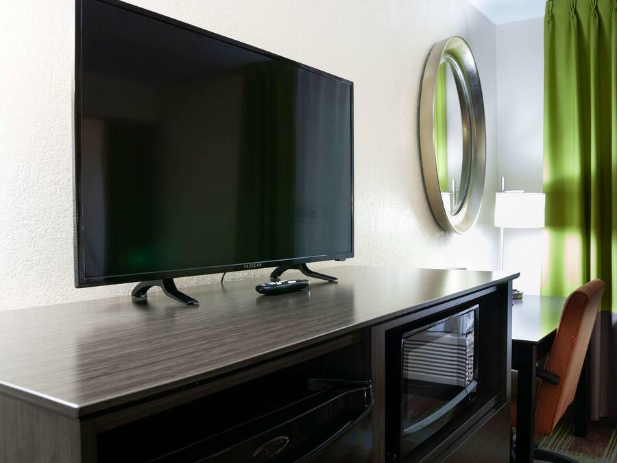 A modern hotel room with a flat-screen TV on a wooden console, a round mirror, a green curtain, and a chair.