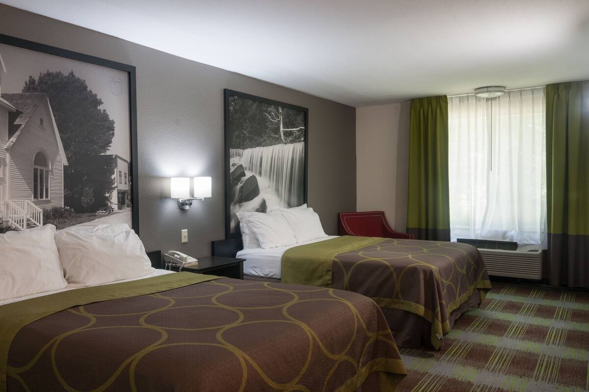 A hotel room with two beds, a nightstand, a red chair, a window with curtains, and a wall-mounted phone.