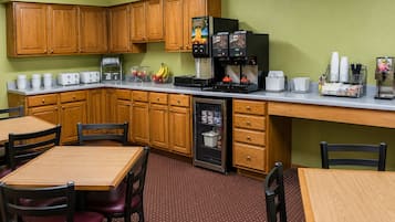 A compact kitchen with wooden cabinets, a refrigerator, a coffee machine, and a microwave. There are two wooden tables with chairs, and a small refrigerator with a freezer drawer.