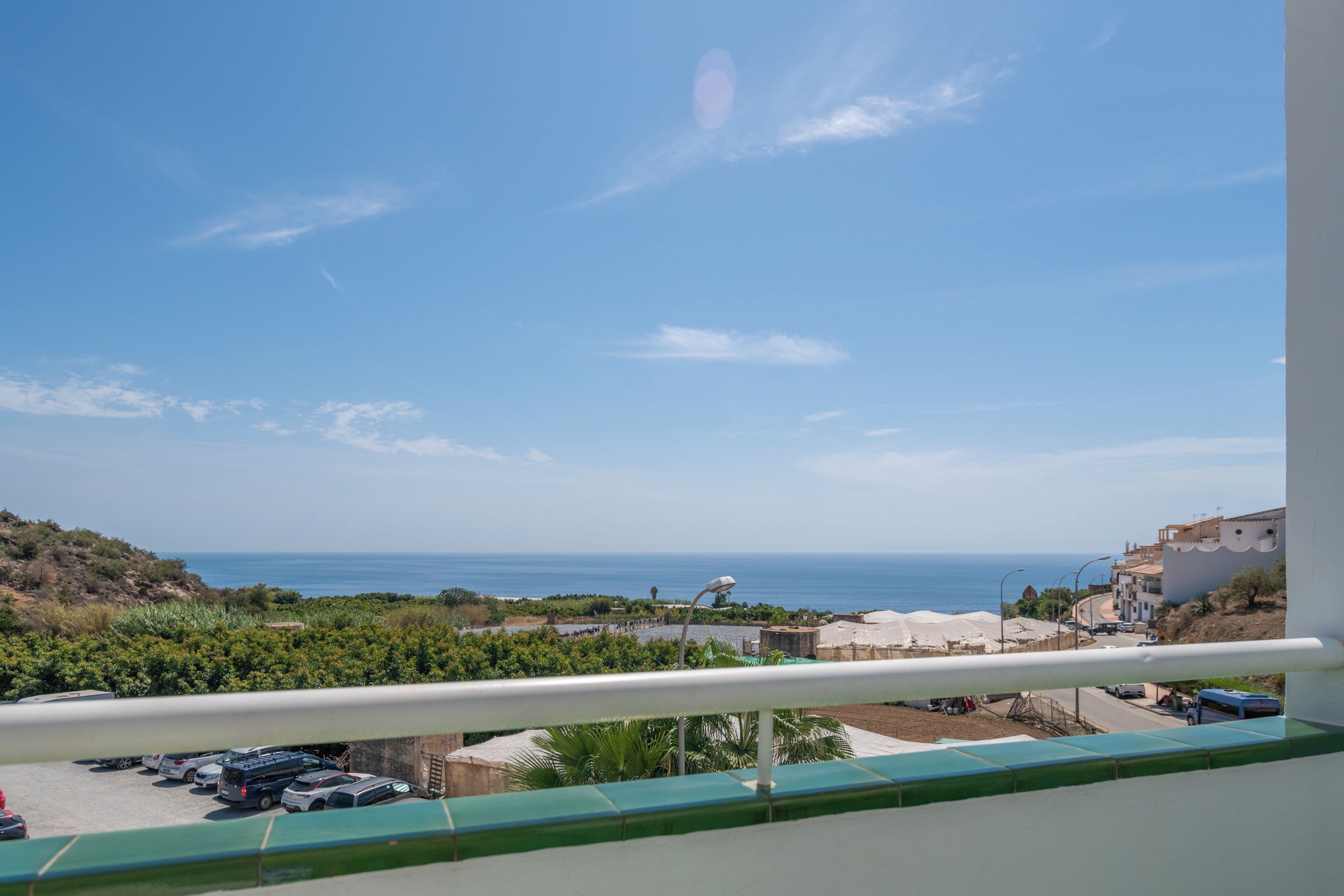 Un balcón con vista al mar, coches aparcados y edificios a lo lejos.