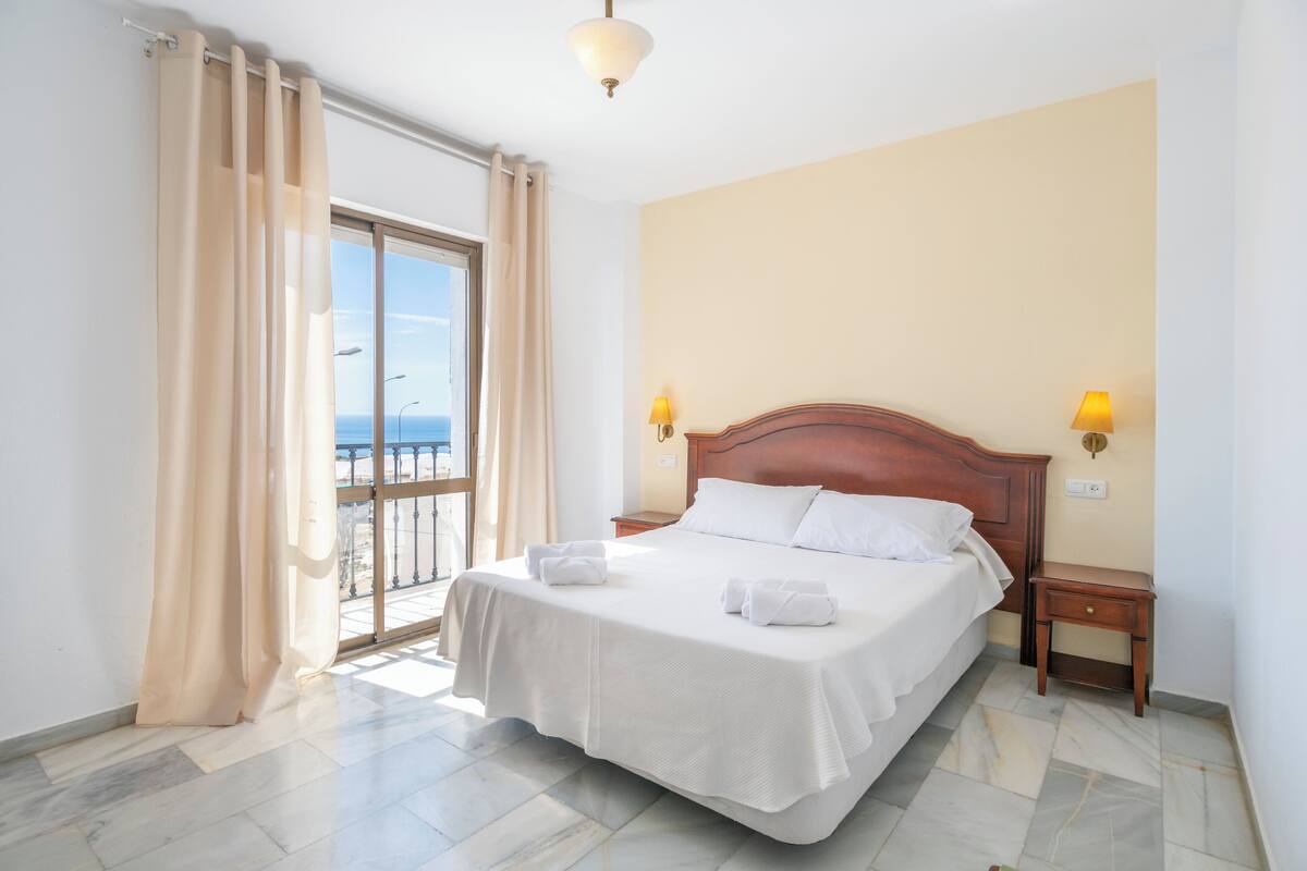 Une chambre d’hôtel avec un grand lit, deux lampes de chevet, une tête de lit en bois, une petite table de chevet et une vue sur la mer par une porte coulissante.