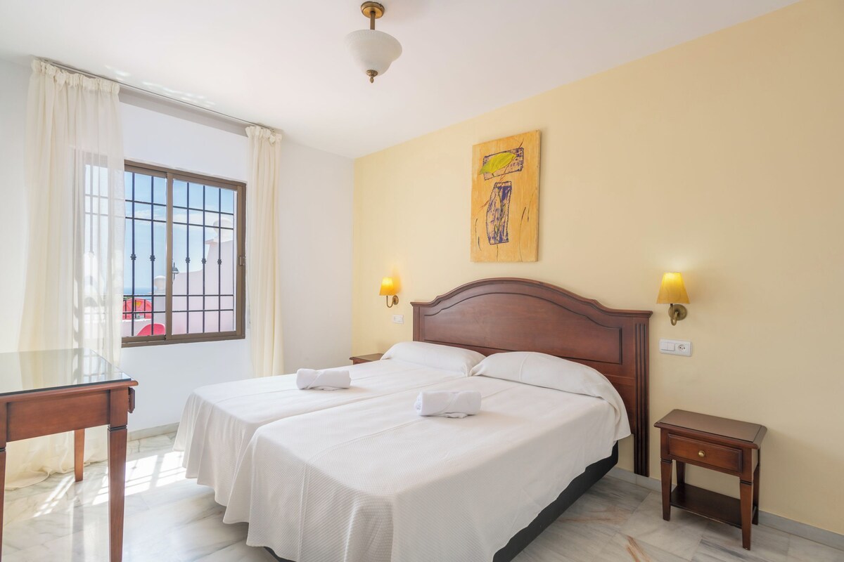 A hotel room with a double bed, two bedside tables, a small desk, a window with curtains, and a wall-mounted artwork.