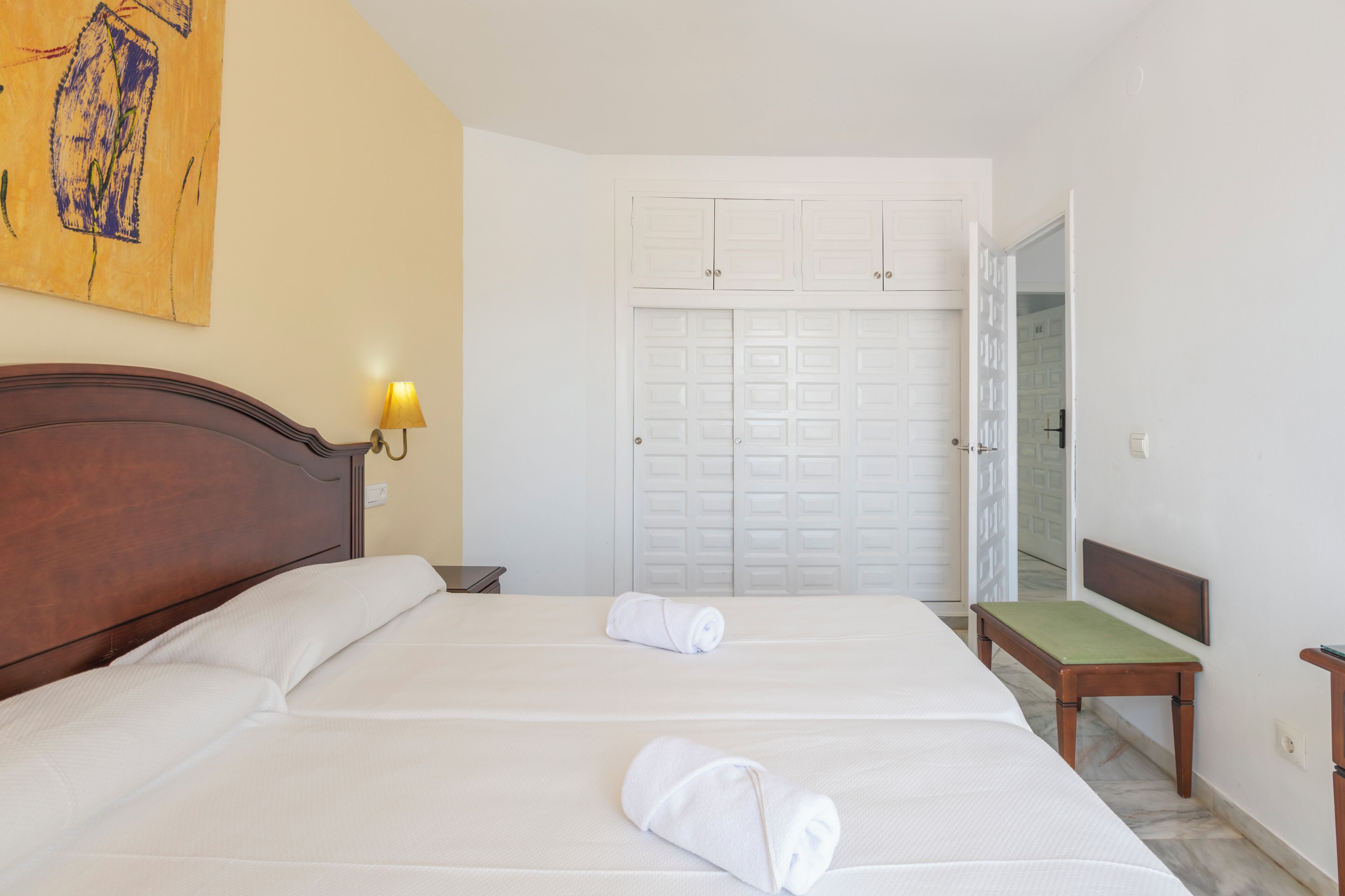 Habitación de hotel con dos camas, cabecero de madera, un banco, una lámpara de pared y una puerta con diseño geométrico.