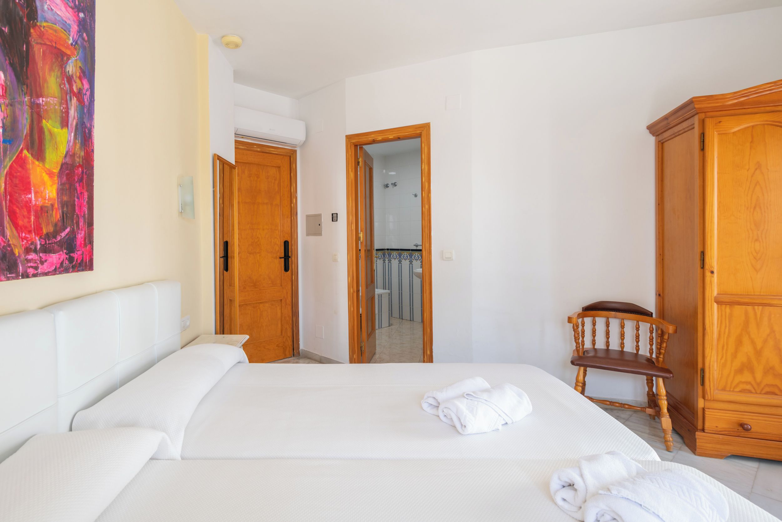 Habitación de hotel con cama, armario de madera, una silla y baño visible a través de una puerta abierta.