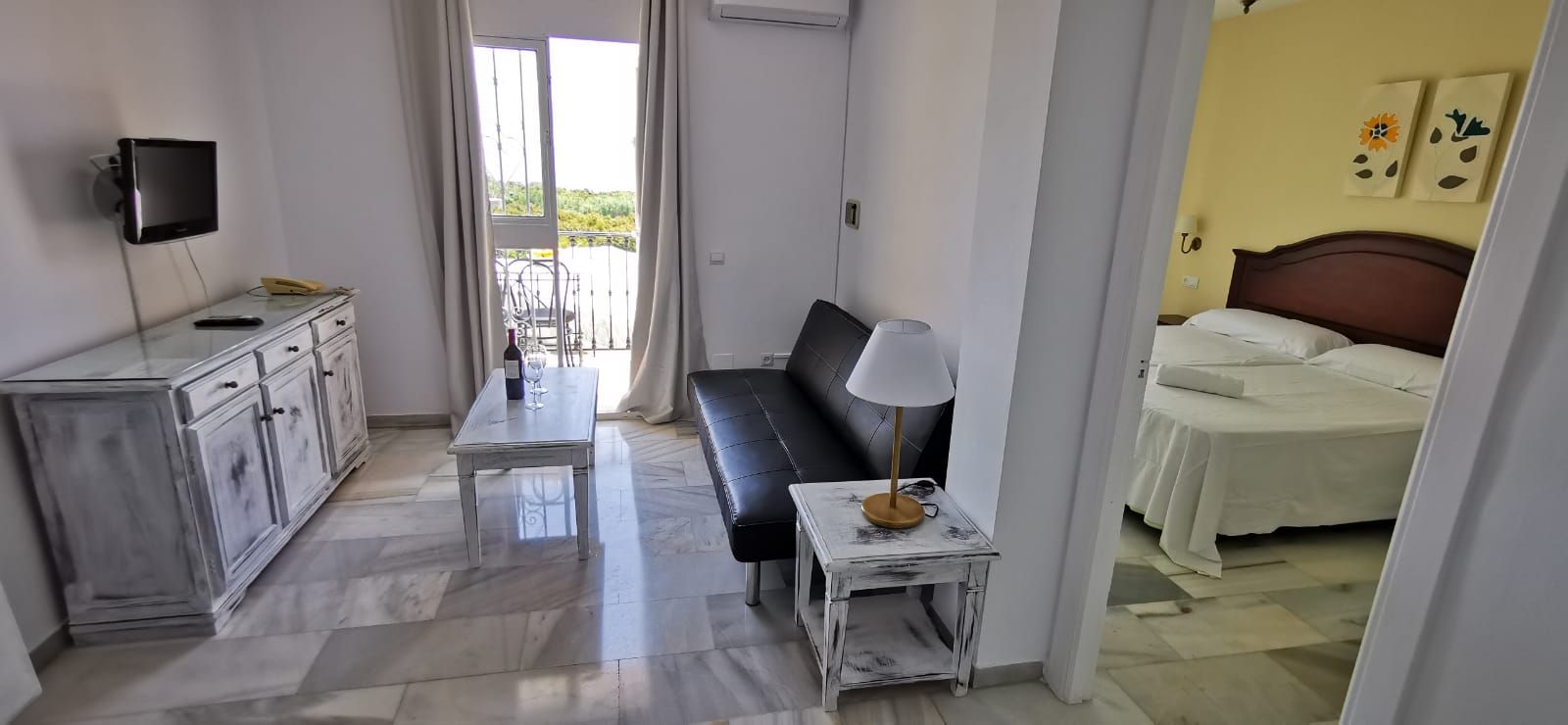 A hotel room with a bed, a desk with a chair, a small table with a lamp, a TV mounted on the wall, and a balcony with a view.