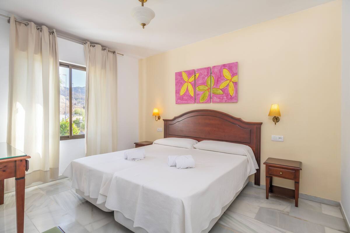 Une chambre d’hôtel avec un grand lit, deux tables de chevet, une commode, une fenêtre avec des rideaux et un mur orné d’un tableau aux couleurs rose et jaune.