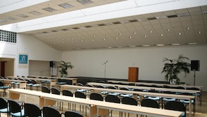 Une salle de conférence avec des rangées de tables et de chaises, un pupitre et deux plantes en pot.