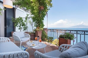 A balcony with white wicker furniture, a table with plates and a drink, potted plants, and a view of the sea and a mountain.