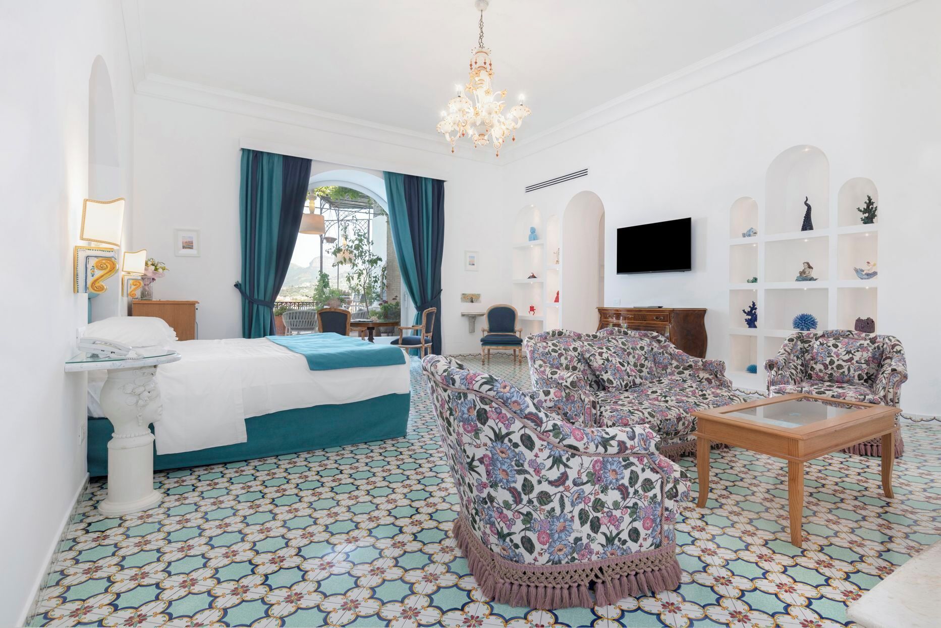 Une chambre d’hôtel avec un lit, deux fauteuils à motifs floraux, une table basse en bois, un téléviseur et une bibliothèque.