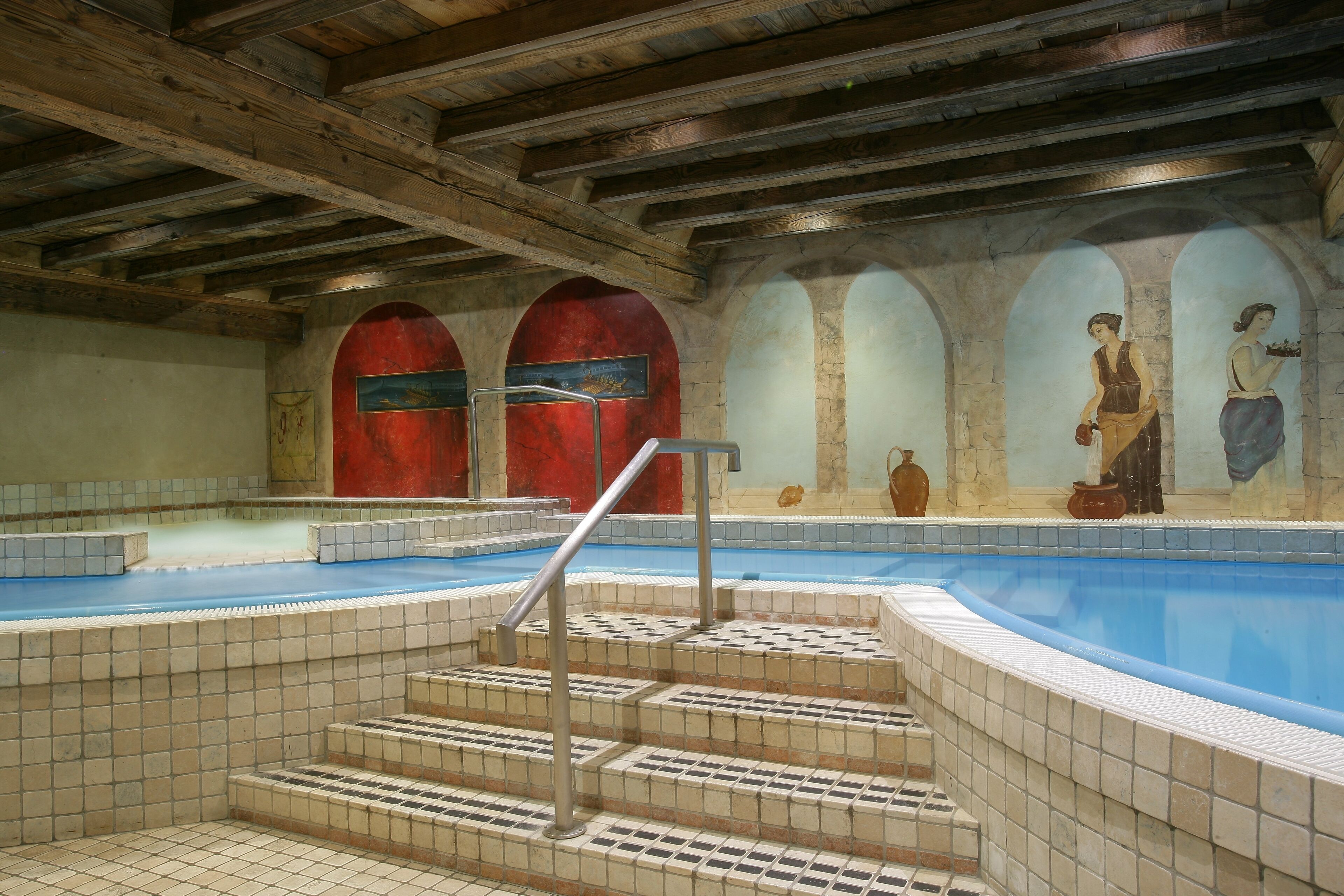 An indoor swimming pool with tiled steps leading to the water, wooden beams on the ceiling, and murals on the walls.