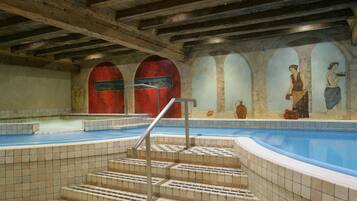 An indoor swimming pool with tiled steps leading to the water, wooden beams on the ceiling, and murals on the walls.
