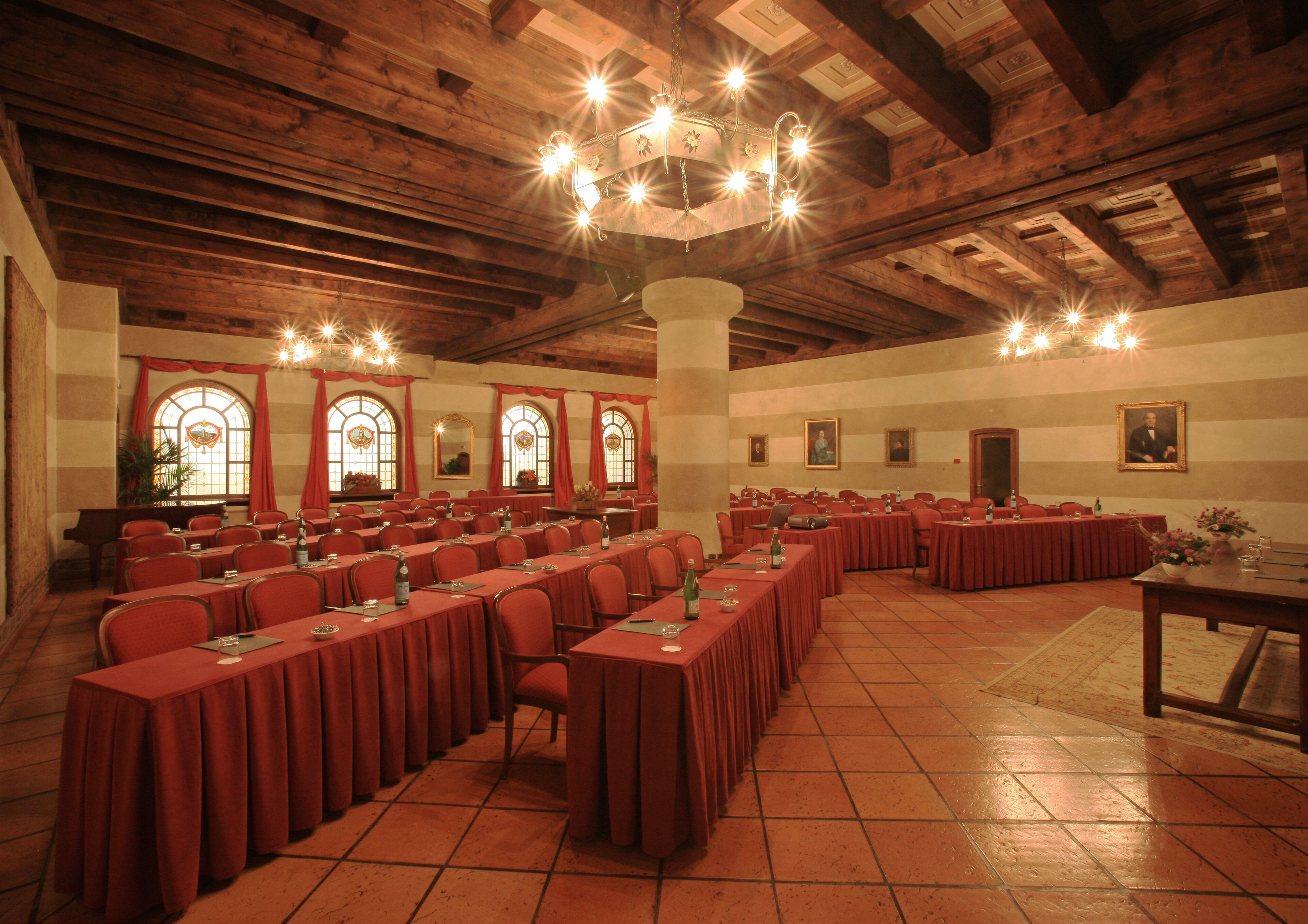 A large conference room with multiple tables and chairs, wooden ceiling beams, and large windows with red curtains.
