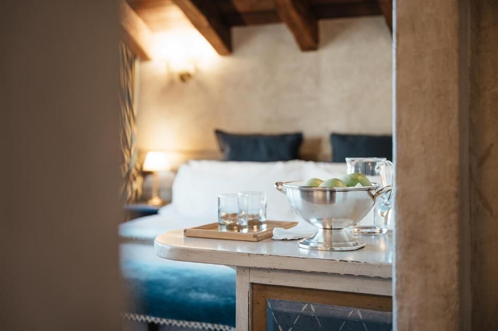 A bed with pillows, a bedside table with a bowl of apples and glasses, and a lamp.