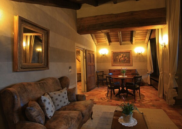 A cozy living room with a brown leather sofa, a dining table with chairs, a large framed painting, and a wooden ceiling.