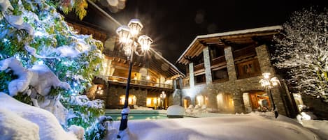 Un espacio exterior cubierto de nieve con un edificio de paredes de piedra y vigas de madera, iluminado por luces cálidas.