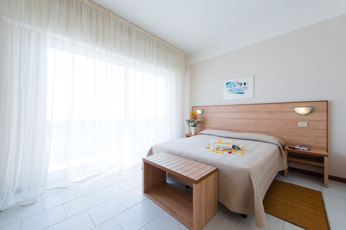 A hotel room with a bed, bedside tables, a wooden bench, a window with sheer curtains, and a framed picture on the wall.