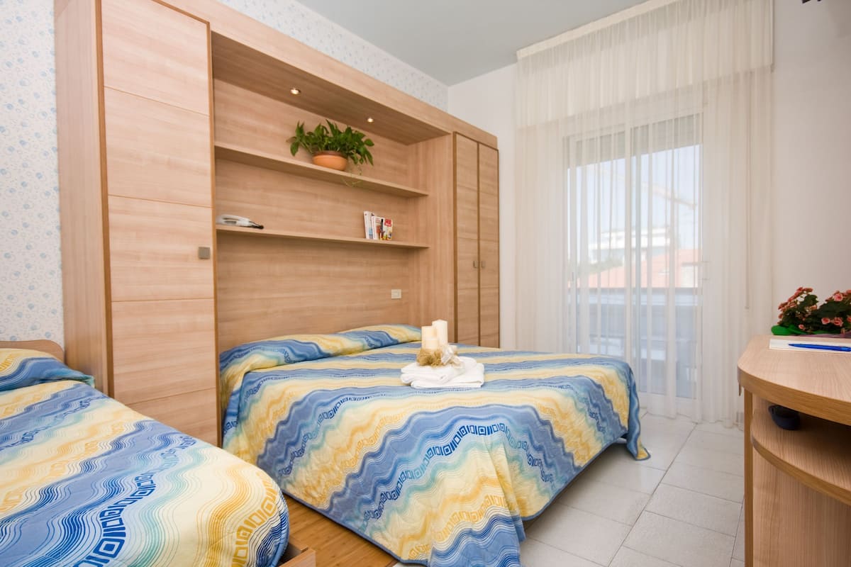 A bedroom with two beds, wooden wardrobe, a small table with a plant, and a window with sheer curtains.